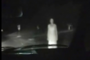 A ghostly woman stands in front of a car, point of view from the passenger's side.